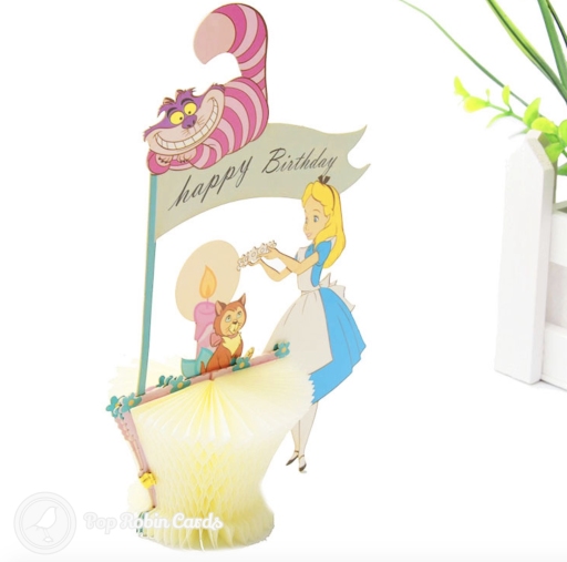 Pop-up of Alice in Wonderland in a blue dress, with the grinning Cheshire Cat above a happy Birthday flag and a pink candle.