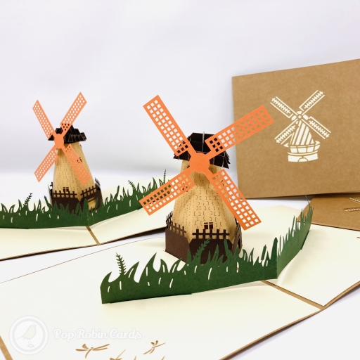 Pop-up Dutch windmill with orange lattice sails and a tan tower behind dark green paper grass, beside a brown cover