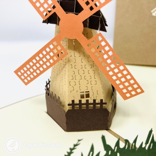 Pop-up Dutch windmill with orange lattice sails and a tan tower behind dark green paper grass, beside a brown cover