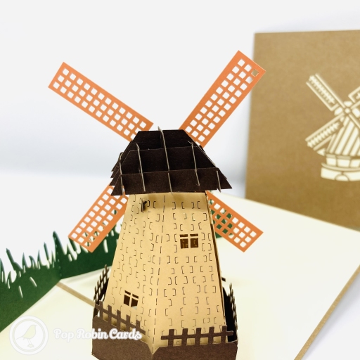 Pop-up Dutch windmill with orange lattice sails and a tan tower behind dark green paper grass, beside a brown cover