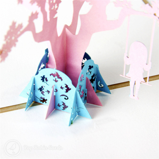 Pop-up pink tree with leaf cut-outs, a blue girl on a swing hanging from a branch, and a blue leaf-patterned base.