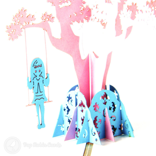Pop-up pink tree with leaf cut-outs, a blue girl on a swing hanging from a branch, and a blue leaf-patterned base.
