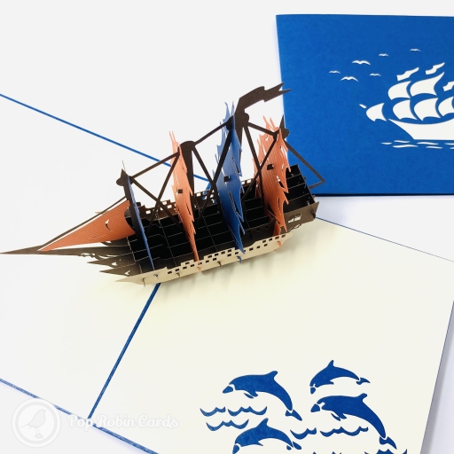 A pop-up galleon with orange and blue sails on a brown hull, beside a blue cover and four blue dolphins.