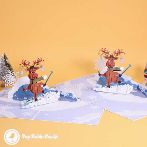 Rudolf the reindeer skiing in a 3D pop-up card, in a striped scarf with lit antlers, on a snowy slope with paper snowflakes