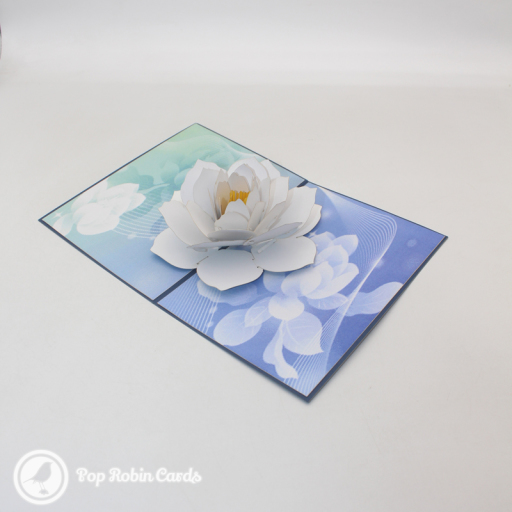 White paper lotus with a yellow centre pops up from a blue and green card printed with faint lotus flowers