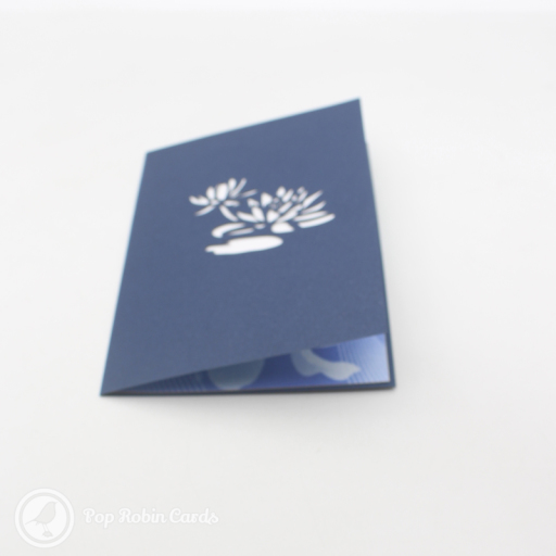 White paper lotus with a yellow centre pops up from a blue and green card printed with faint lotus flowers